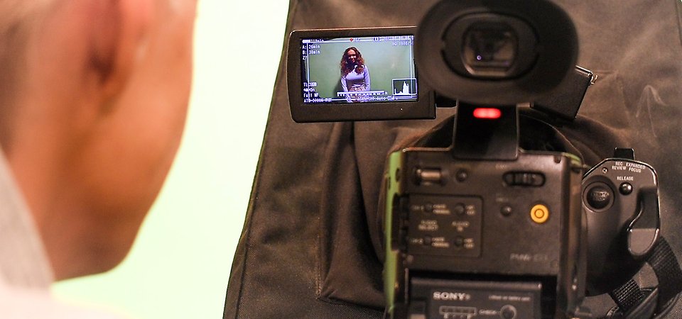 En filmkamera. På skärmen syns en tjej såt framför en green screen.